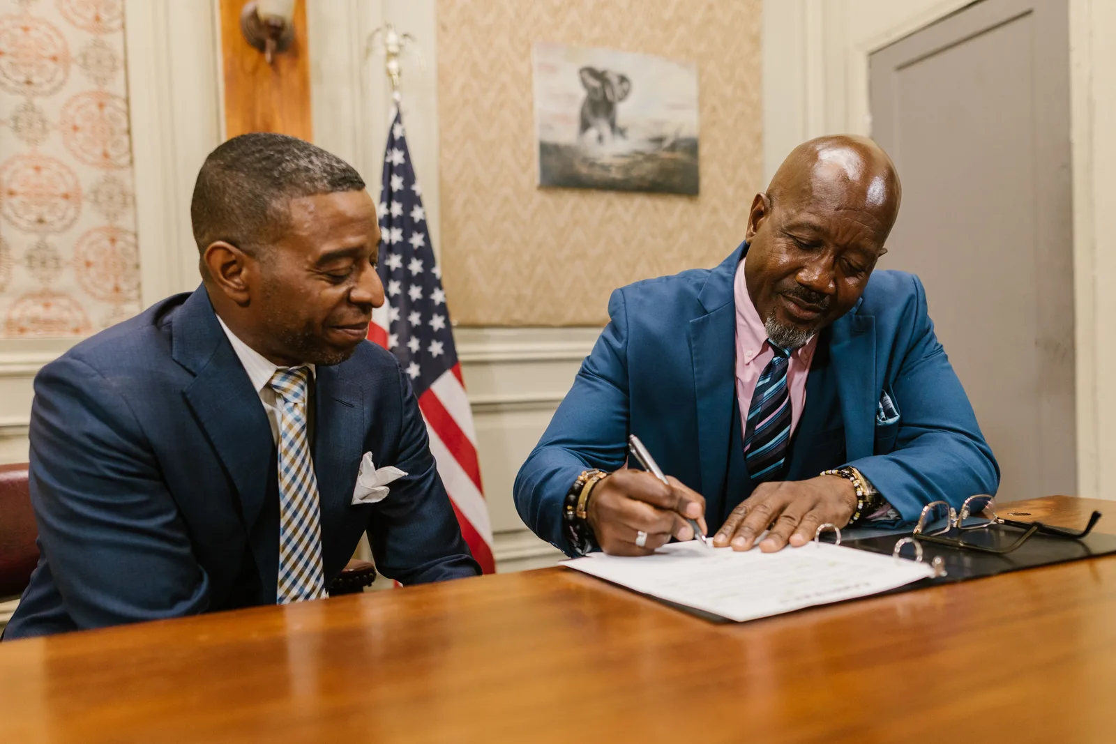 Two businessmen in suits signing documents in an office setting with an American flag. A code of conduct only works if it survives real pressure, not just launch day